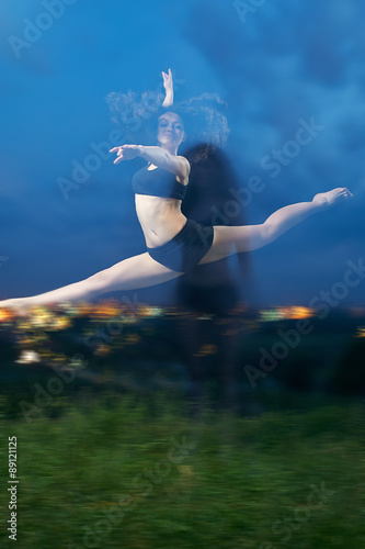 pretty, young, smiling dancer performs splits in the air at the grassy field in front of the night lights of the city.
