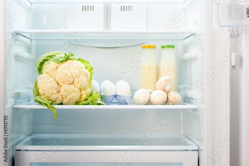 Cauliflower, white eggs, champignon mushrooms and two glass bottles of yoghurt on shelf of open empty refrigerator. Weight loss diet concept.