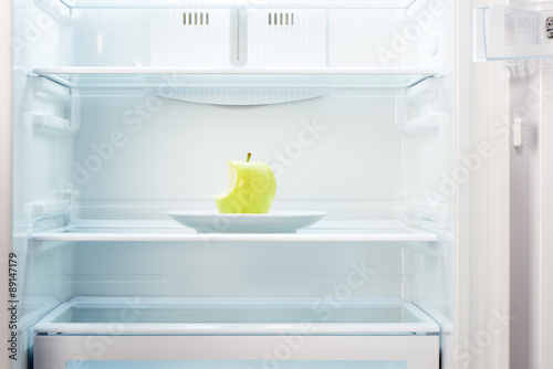 Green bitten apple on white plate in open empty refrigerator. Weight loss diet concept.