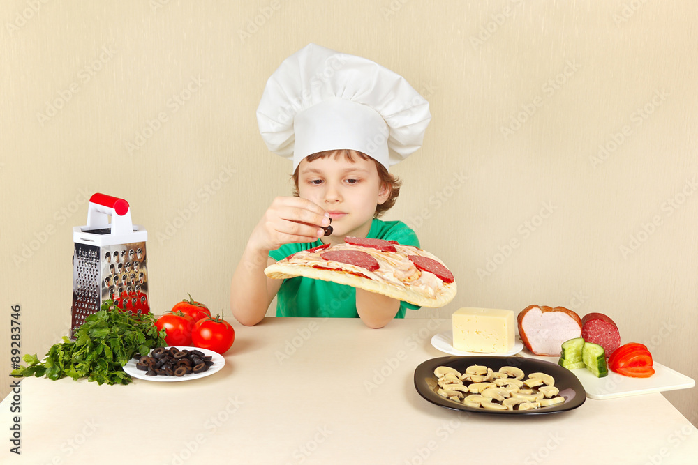 Little boy in chefs hat puts olives on the pizza crust Little boy in chefs hat puts olives on the pizza crust