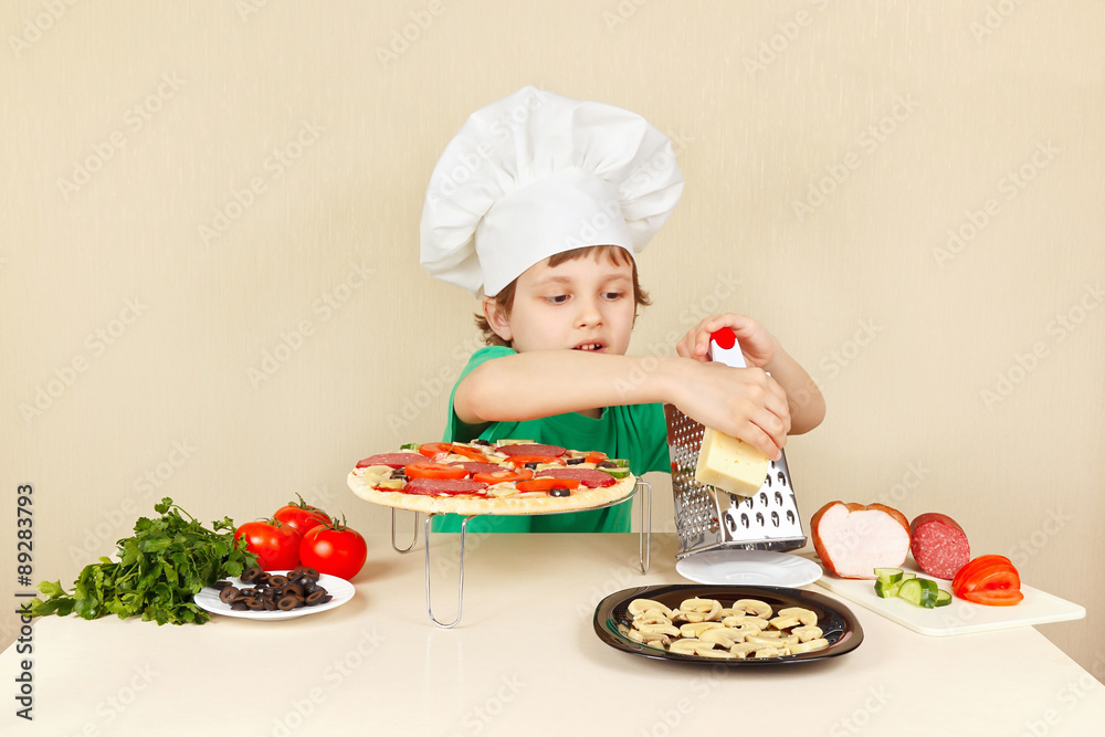 Little funny boy in chefs hat rubs on a grater cheese for pizza Little funny boy in chefs hat rubs on a grater cheese for pizza