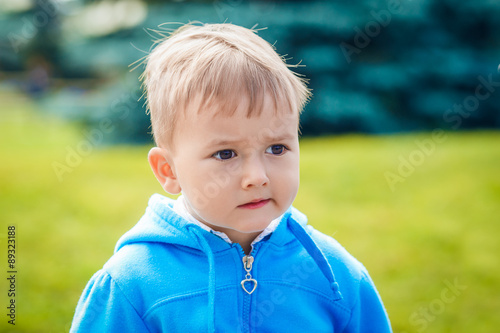 Closeup portrait of cute Caucasian boy with blond hair and dark brown eyes with funny face expression in blue hoodie outside in park on summer day, backlit with sun, rim light of his figure