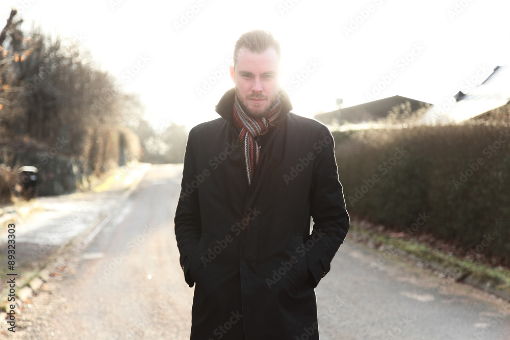Handsome man in his 20s, standing outside on a cold winter day wearing a black jacket and scarf looking straight into camera. Pensive with the sun straight behind him. Handsome man in his 20s, standing outside on a cold winter day wearing a black jacket and scarf looking straight into camera. Pensive with the sun straight behind him.