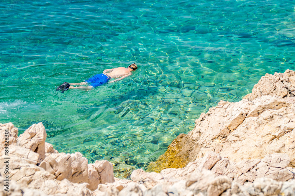 A man snorkeling in a crytsla clear blue sea or ocean by the rocky coast