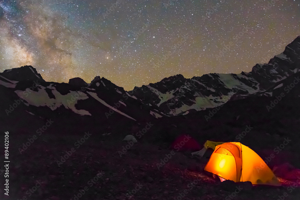 Night mountain landscape with illuminated tent.
Silhouettes of snowy mountain peaks and edges night sky with many stars and milky way on background illuminated orange tent on foreground