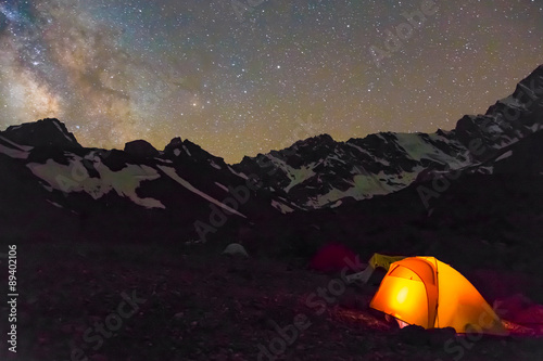 Night mountain landscape with illuminated tent. Silhouettes of snowy mountain peaks and edges night sky with many stars and milky way on background illuminated orange tent on foreground