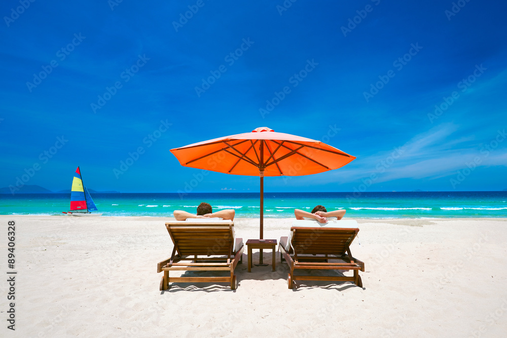 Couple on a tropical beach on deck chairs under a red umbrella.
