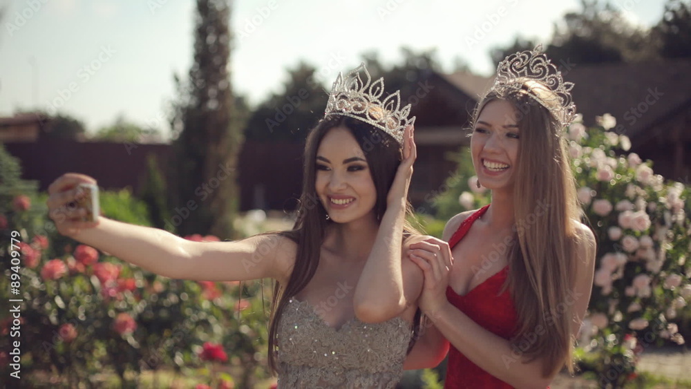Two girls in evening gowns and crowns make selfie on phone in a Two girls in evening gowns and crowns make selfie on phone in a