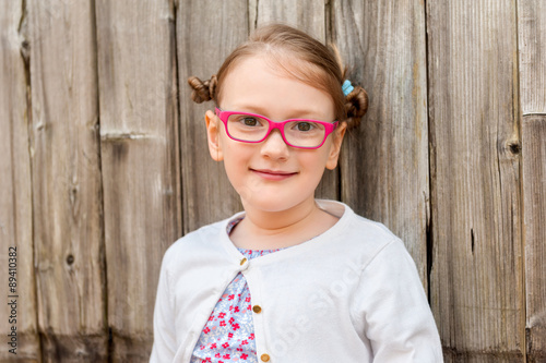 Close up portrait of a cute little girl of 7 years old, wearing pink eyeglasses, standing against wooden background