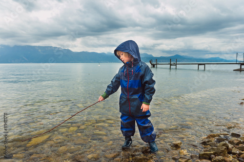 Cute little boy playing by the lake, pretending fishing, wearing blue waterpoof all-in-one suit and rain boots