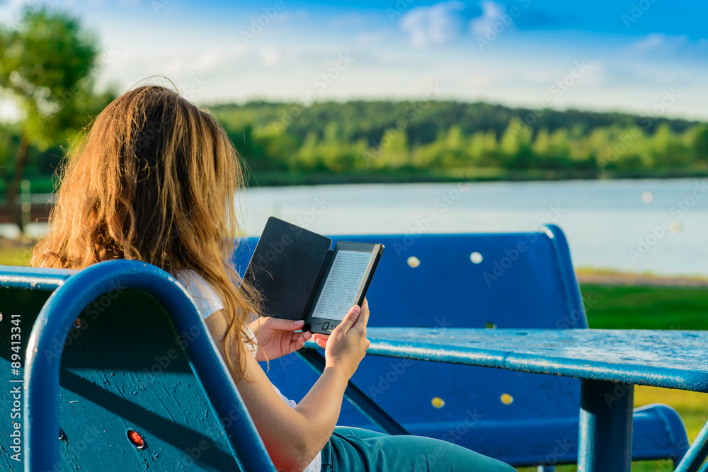 Girl reading from a tablet on the resting-place against a lake.