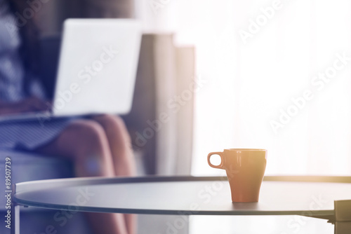 A cup of coffee with Young woman using laptop at home, selective focus on cup of coffee with sunrise streaming in through window and creating flare into the lens.