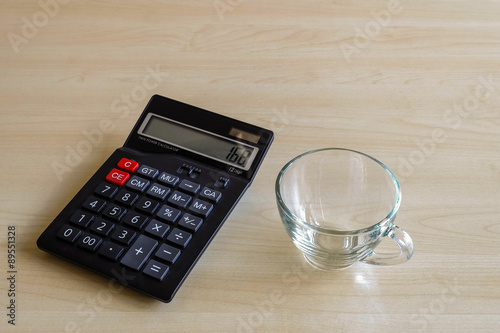 A glass cup and black calculator on wood background under window light