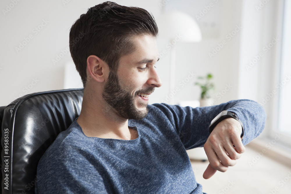 Smiling young man sitting on leather chair looking at his smartwatch Smiling young man sitting on leather chair looking at his smartwatch
