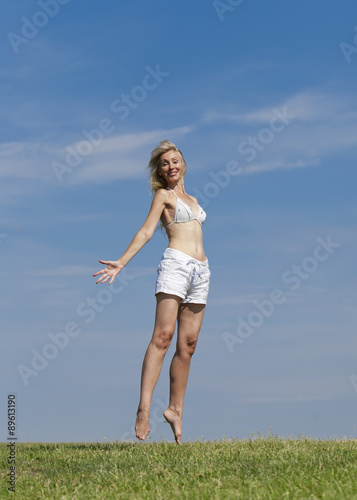 The happy woman in white bikini and shorts jumps in a summer green field against the blue sky..