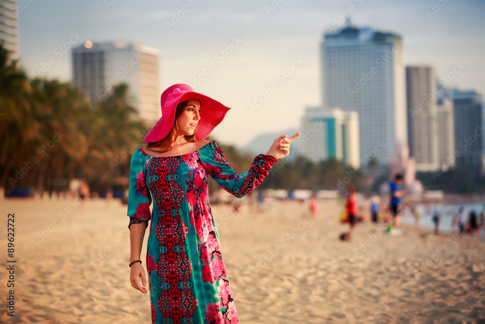 slim girl in long and big hat on beach against city sea