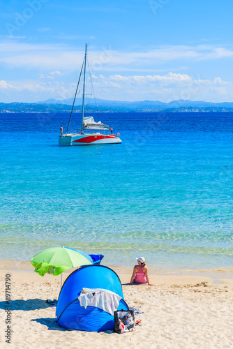 Woman sitting on sandy Grande Sperone beach with catamaran boat on azure sea in background, Corsica island, France