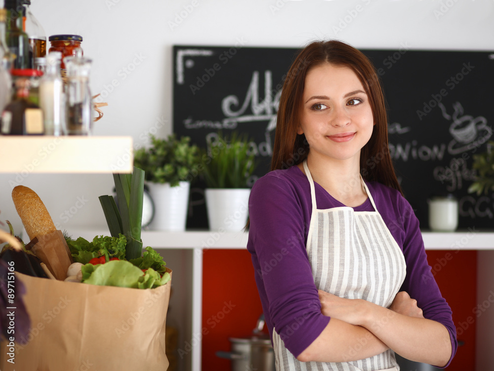 Young woman standing in her kitchen near desk with shopping bags Young woman standing in her kitchen near desk with shopping bags