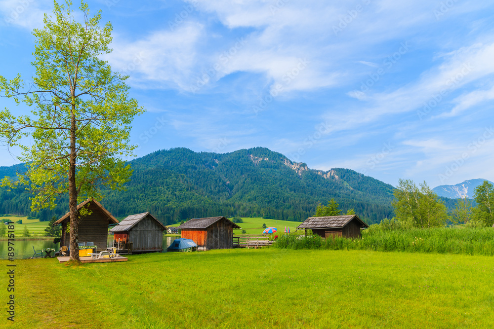 Green meadow in small alpine village on shore of Weissensee lake with traditional boat houses in background, Austria