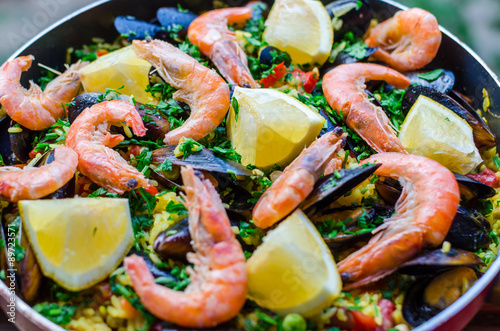 Close up fresh juicy prepared classic spanish dish paella with seafood, blueshell mussels, shrimps, rice with saffron spice, vegetables, smoked sausages and peaces of lemon with parsley in frying pan