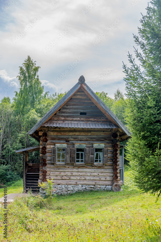 Old historic peasant's log house in rural Russia, Siberia surrounded by summer forest and wildflowers under misty clouds.