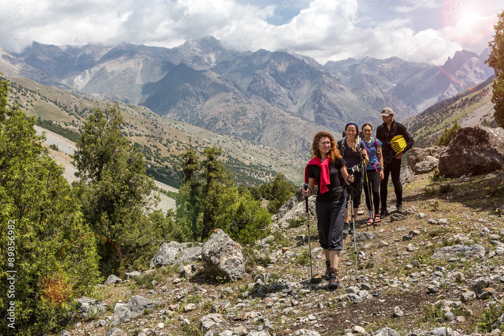 Group of people walking on trail Men and women going up with backpack luggage and hiking gear on bright mountain landscape background with sun rising and high peaks behind