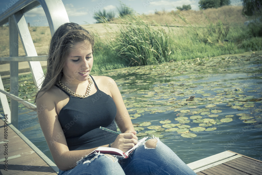 Adolescente de 15 años escribiendo en el embarcadero. Chica rubia adolescente escribiendo en su diario junto al rio. Adolescente de 15 años escribiendo en el embarcadero. Chica rubia adolescente escribiendo en su diario junto al rio.