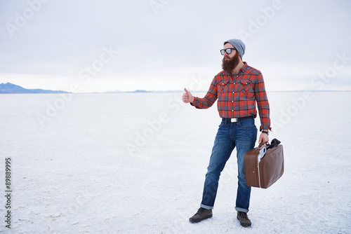 nonchalant entitled bearded hipster hitch hiker thumbing for a ride in empty vast salt flats with retro suitcase