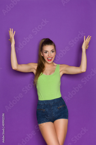 Excited Young Woman. Beautiful young woman in lime green shirt and jeans shorts standing with arms raised and shouting. Three quarter length studio shot on violet background.