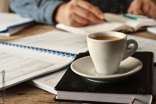 In the foreground is a cup of coffee on the diary for records closeup. In the background, a man working in the office. The concept of long hours of work. Copy space. Free space for text