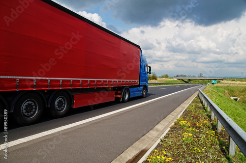 Red truck on asphalt highway. Concrete bridge over the highway in the distance. Meadow with flowering dandelions. Sunny day with blue sky and clouds.