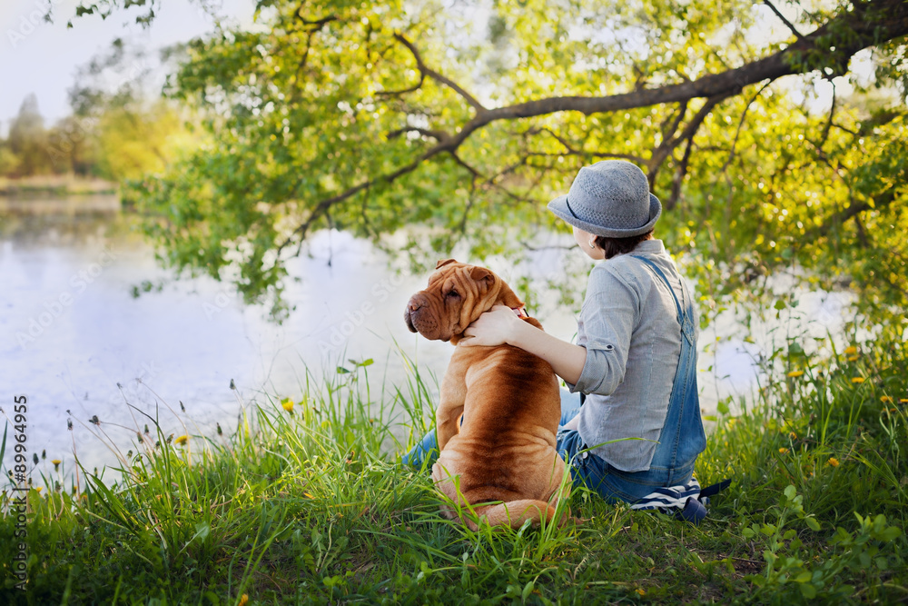 young woman in a hat with dog Shar Pei sitting in the field and looking to the river in golden sunset light, true friends forever, people concept, meditation and relaxation, back to camera