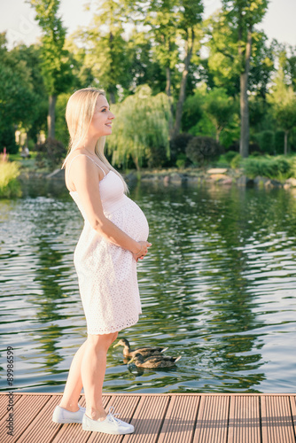 Pregnant woman in white dress holding belly near lake.