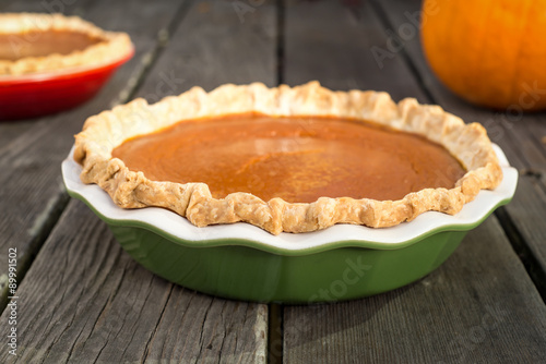 Freshly baked pumpkin pies cooling on rough wood boards. Green and red ceramic pie plates. Fresh pumpkin in the background.