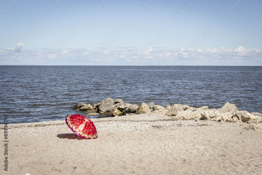 horizontal image of a red patterned umbrella lying on a sandy beach beside the ocean
with large rocks jutting out under a clear blue sky in the summer time horizontal image of a red patterned umbrella lying on a sandy beach beside the ocean
with large rocks jutting out under a clear blue sky in the summer time