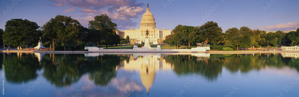 This is the U.S. Capitol showing the West side view at sunset. The Capitol is in front of the reflecting pool, showing a reflection of itself in the pool. The Capitol is surrounded by green trees and a blue sky with a public statue in front of it.