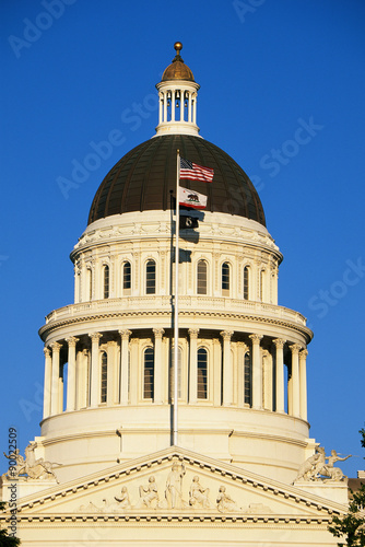 This is the State Capitol dome at sunset. It has several flags waving in the wind on a flagpole in front of it, the top one is the American flag.