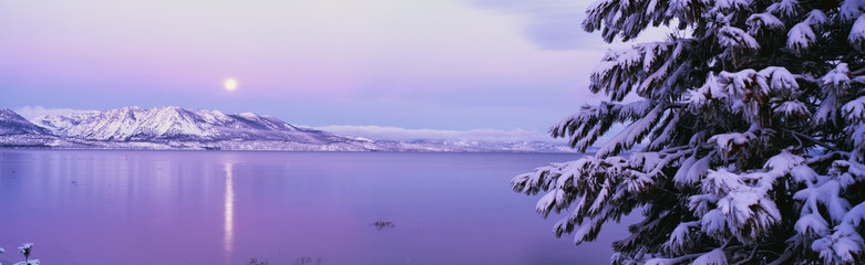 This is Lake Tahoe after a winter snow storm. There is a full moon rising over the lake and the trees and mountains are covered in snow.