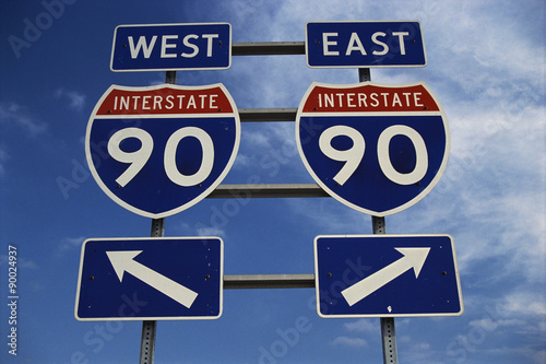 This is a road sign on the New York State Freeway. It points out the direction for Route 90 to go east or west. The signs are blue, red and white against a blue sky.