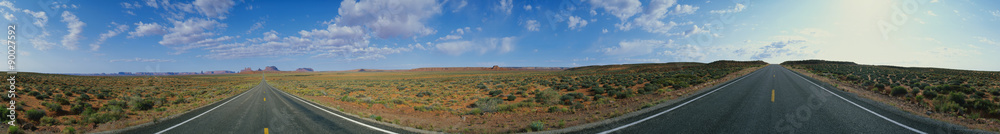 This is a 360 degree image of a fork in the road. The rocks of Monument Valley are small in the background and there is desert foliage on all sides of each road. The sky is blue with white puffy clouds.