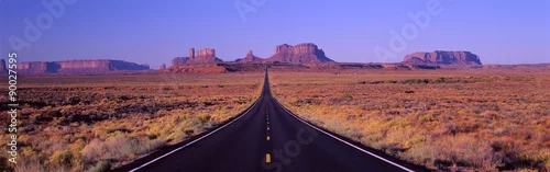 Obraz To jest trasa 163, która biegnie przez rezerwat Indian Navajo. Droga biegnie w górę środka i staje się mniejsza w nieskończoność. W tle znajdują się czerwone skały Monument Valley. Pustynne rośliny zarośla znajdują się po obu stronach drogi.