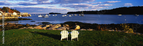 This is an image of two white lawn chairs facing toward the nearby harbor. There are fishing boats moored in the harbor in this small lobster village. The chairs are surrounded by green grass and the harbor has clear blue water. The sky is blue with white clouds.