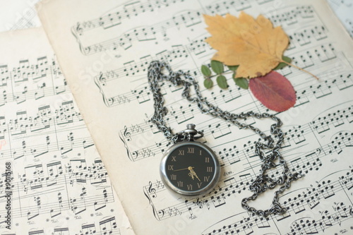 Old watch and dead tree leaves laying on old piano musical notes