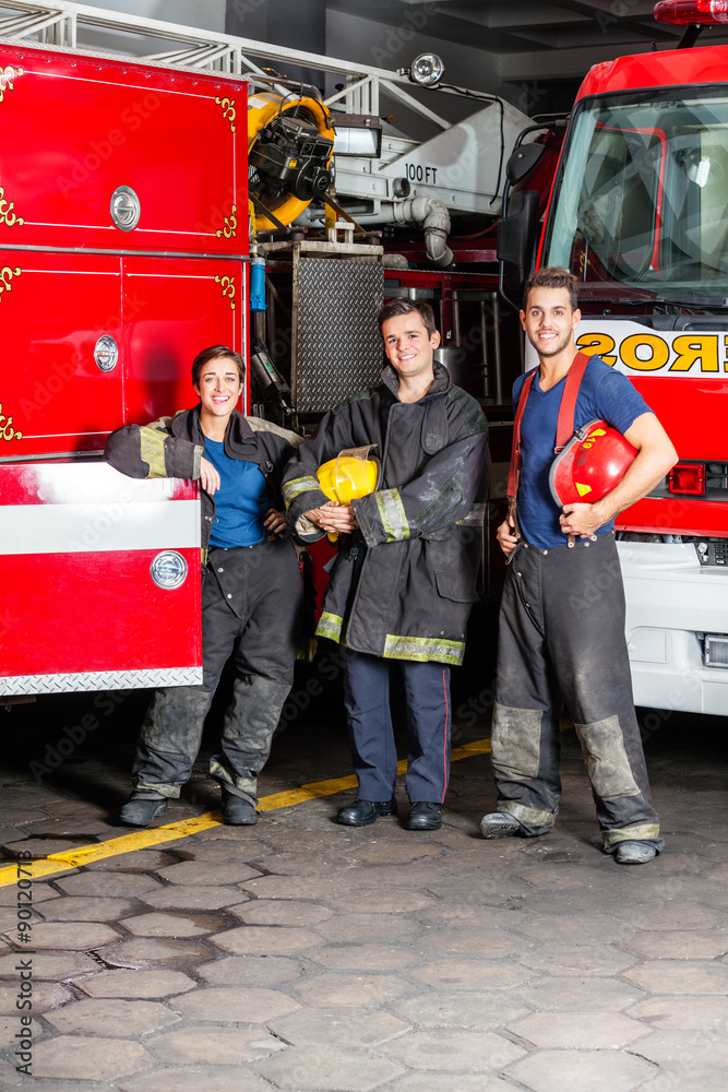 Fototapeta premium Happy Young Firefighters Standing Against Trucks
