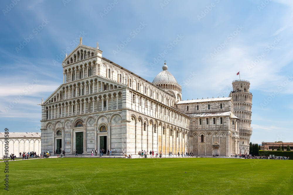 Beautiful view of the Cathedral and the Tower of Pisa. Italy. The Cathedral and the Tower are on the background of blue sky with light clouds. In the foreground a beautiful green lawn. Sunny day.