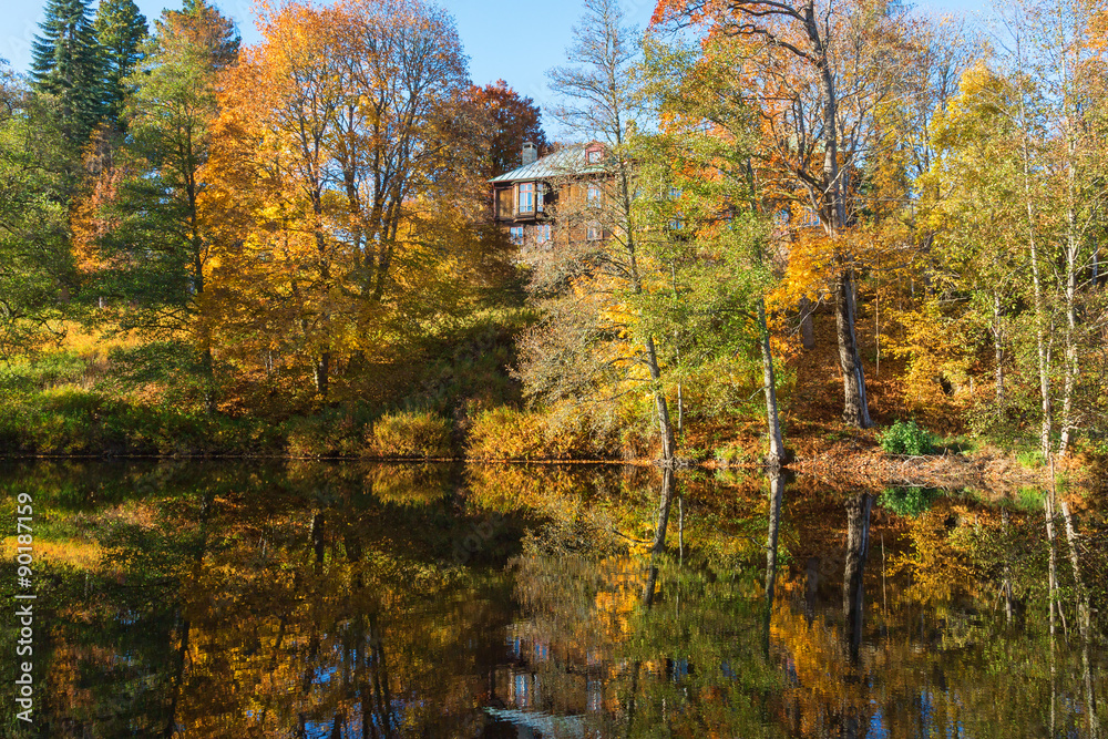 Deciduous forest in autumn colors reflecting in the water surface of the lake with a house on the hill