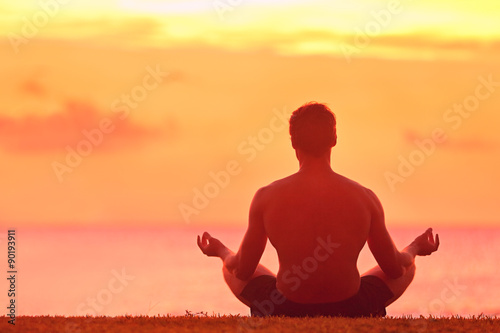 Meditation and Yoga. Rear view of young man meditating in lotus position. Male is practicing yoga at beach. He is doing relaxation exercise at beach during sunset.