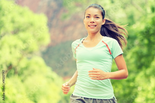 Fototapeta Naklejka Na Ścianę i Meble -  Young woman jogging while smiling. Beautiful mixed race Asian / Caucasian female is in sports clothing. Attractive lady is in park.