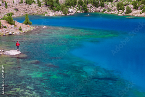 Mountain Lake and Alone Traveler Vivid Green Blue Azure Water Surface of Lake with Transparent Pure Water and Sones Visible on Bottom Body Hiker Staying on Rock Among