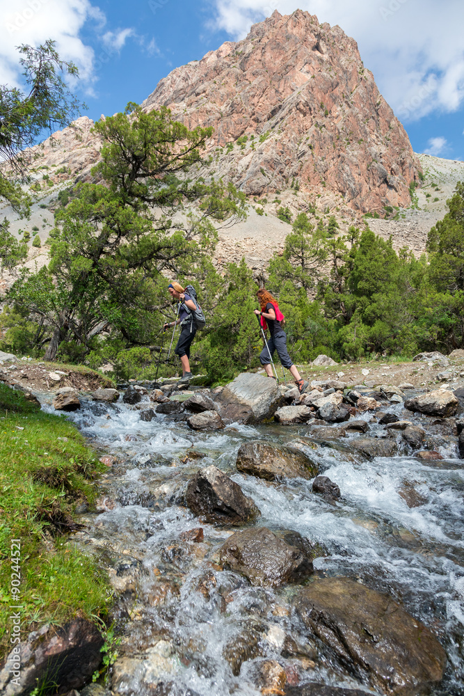 Two hikers crossing fast flowing river People going across mountain creek with fast streaming water jumping on the rocks green meadow and forest along river forest wild landscape and sun on background